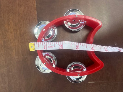 Red tambourine with silver jingles on a wooden surface, accompanied by a measuring tape.