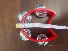 Red tambourine with silver jingles on a wooden surface, accompanied by a measuring tape.