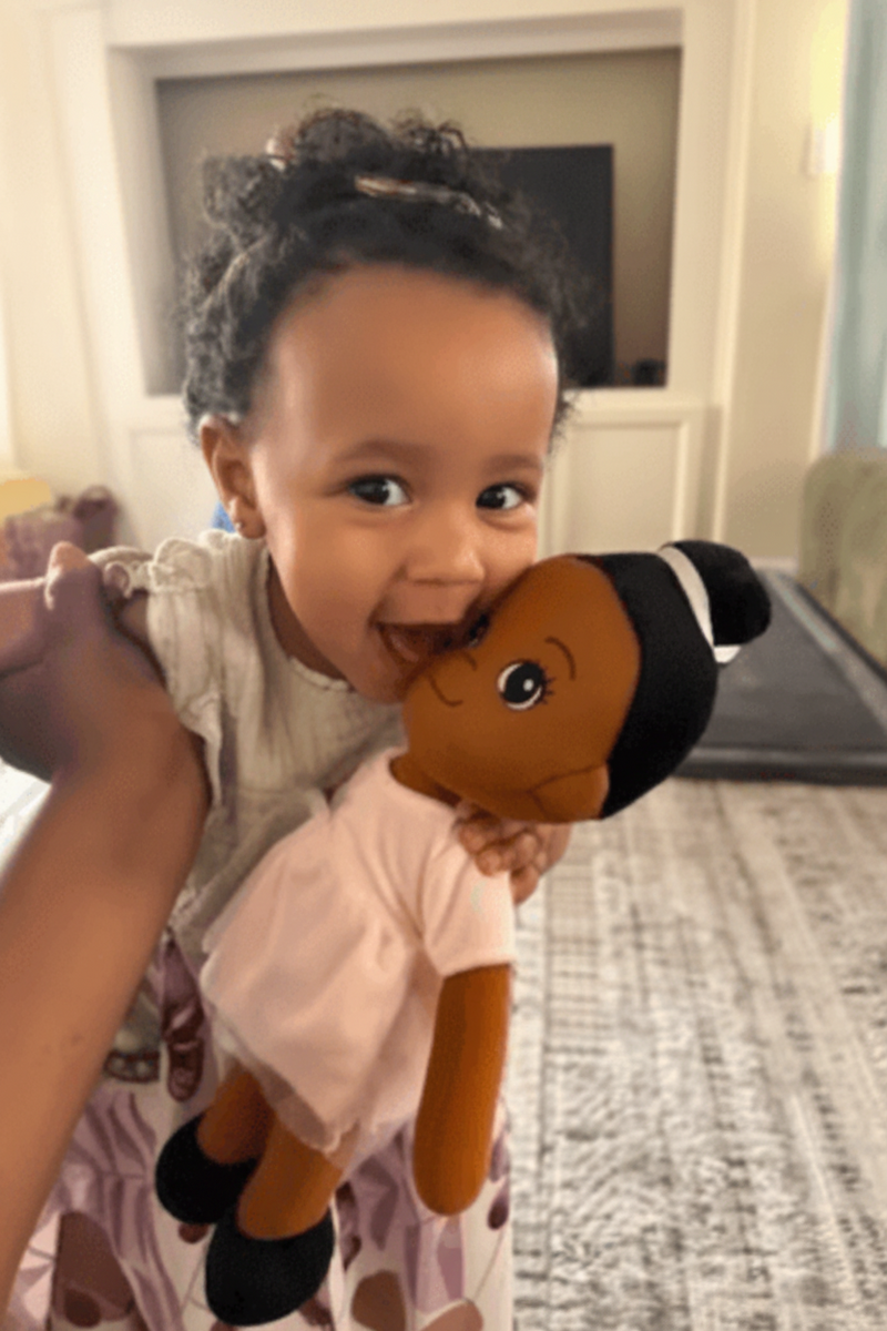 A young child holding a small doll with dark skin, black hair, and a light pink dress in a home setting.