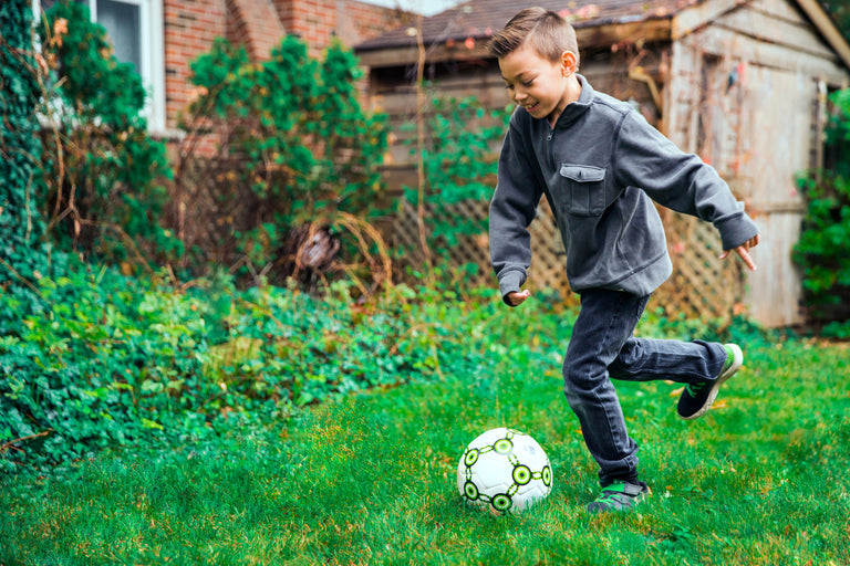 A young boy in a gray jacket plays with a soccer ball on a grassy lawn in front of a wooden shed.